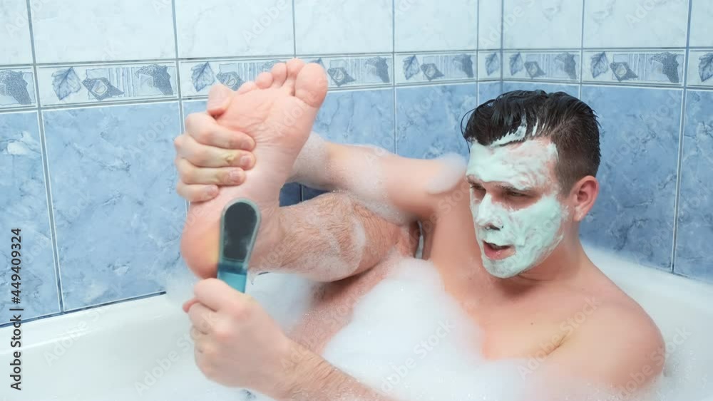 Young man with mask on his face is peeling his foot sitting in bathroom with foam at home. Spa beauty treatment. Man wellness, skincare and self love concept. Relaxing and enjoying his time.