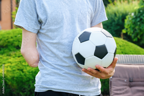 Full length young caucasian man with amputated arm, holds of soccer ball while standing on the lawn in the backyard of his home, disability rehabilitation and active lifestyle © Serhii