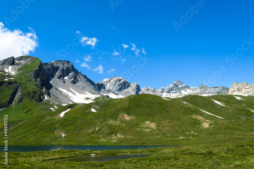 Snowcapped peaks above green pastures in summer. View of a lake at the bottom of the valley with meadows. Mountains of the Italian Alps.
