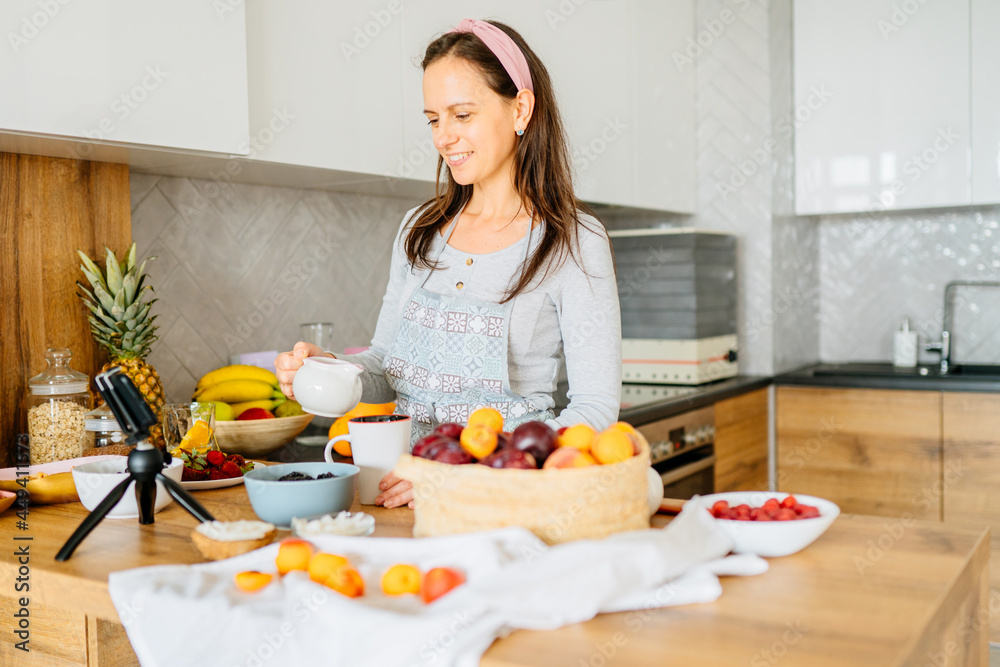Food, culinary bloggers concept. Smiling middle age woman food blogger wearing apron and recording her cooking by mobile phone on tripod in kitchen, gesturing at phone camera, healthy fresh food.