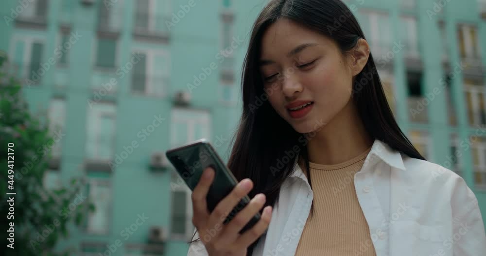 Attractive Asian Girl in White Shirt standing outdoors and looking at Smartphone. Browsing Modern Phone, reading Positive News, or Scrolling Posts. Using apps for chatting. Messenger. Social Media. Attractive Asian Girl in White Shirt standing outdoors and looking at Smartphone. Browsing Modern Phone, reading Positive News, or Scrolling Posts. Using apps for chatting. Messenger. Social Media.