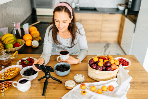 Congratulations holiday at distance  online call  video food blog at home. Happy pretty mature woman gesturing and look at smartphone webcam in kitchen interior with bright fresh fruits and berries.