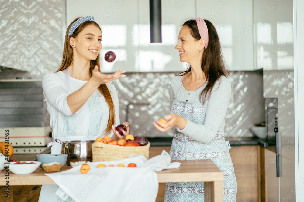 Attractive young adult woman talking with her mature mother throwing violet plum into air in kitchen. Natural healthy snack food - fruit pastille from different berries and fruits. Attractive young adult woman talking with her mature mother throwing violet plum into air in kitchen. Natural healthy snack food - fruit pastille from different berries and fruits.