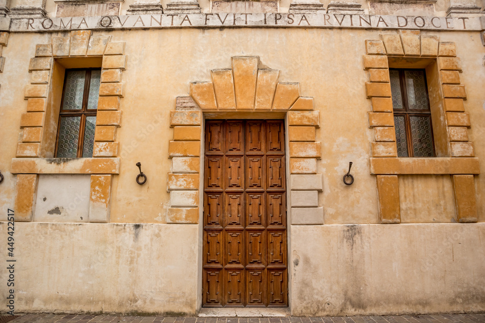 Old building front wall wit wooden door and two windows. Ancient Roman letters caption, warm colors. Eastern Italy