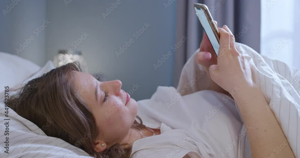 Happy attractive young woman lying in bed, texting on mobile phone. Write messages with smartphone, use apps to communicate.