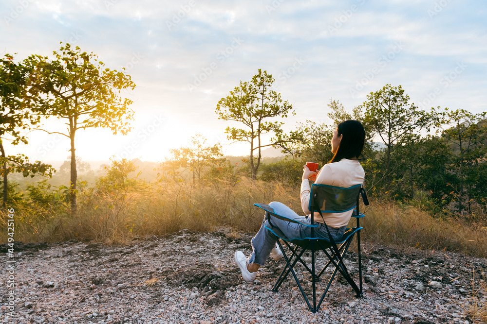 Fototapeta premium Rear view of happy young woman sitting on outdoor chair near a tent in the mountains, drinking coffee with sunrise in morning.