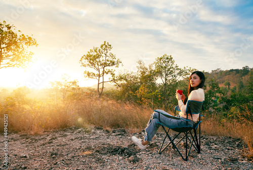 Happy young woman sitting on outdoor chair near a tent in the mountains, drinking coffee with sunrise in morning.