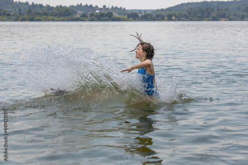 Fototapeta Naklejka Na Ścianę i Meble -  Teenage girl in blue swimsuit bathes in calm cool lake water in hot summer, raising a lot of splashes