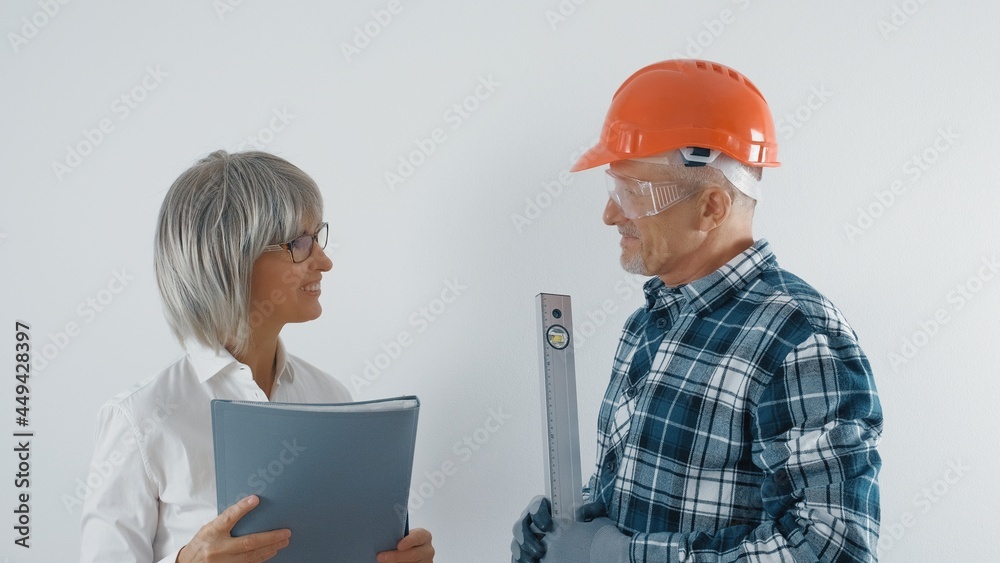An elderly worker in a hard hat and a gray-haired female engineer with a tablet in their hands are discussing plans for the reconstruction of the facility. Making decisions on repairs. An elderly worker in a hard hat and a gray-haired female engineer with a tablet in their hands are discussing plans for the reconstruction of the facility. Making decisions on repairs.
