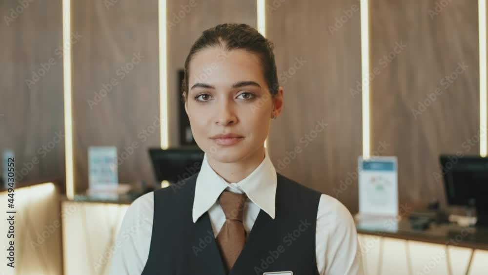 Medium close-up of young attractive hotel manager looking at camera standing at reception of modern high-class hotel. Suitable for deepfake generation or face swap