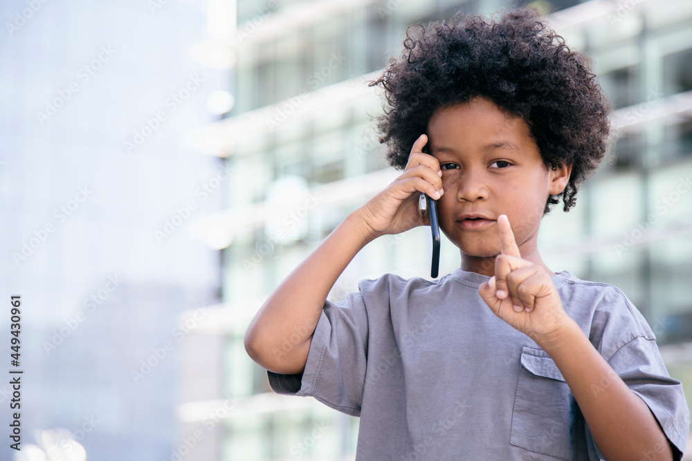 Black boy, with afro hair, talking to his cell phone, raising his finger with a warning expression. With a office buildings background. Children, psychology and smartphones concept. Copyspace