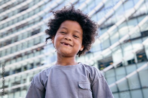 Black boy, with afro hair, looking at the camera. Smiling, missing his two front teeth. With a serious expression. With a office buildings background. Children, future and poverty concept.