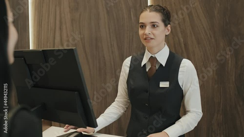 Medium shot of young female receptionist talking politely to guest woman during check-in as looking for available rooms in hotel photo