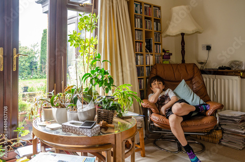 A boy relaxes in a comfy chair with a glass table covered with houseplants in front. A homely lifestyle scene from a family home.