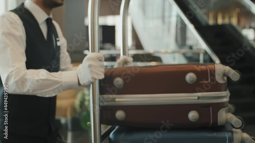Midsection shot of unrecognizable male porter in uniform and gloves carrying baggage cart through modern hotel lobby