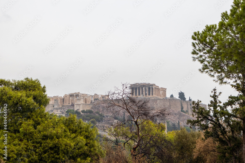 Acropolis hill (Parthenon, Propylaea, Temples, Odeon of Herodes Atticus) view through green trees branches. Athens ancient historical landmark in city center from Filopappou Hill on cloudy day