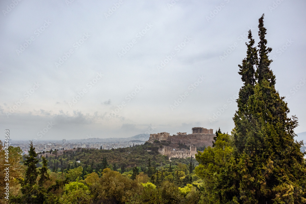 Acropolis hill (Parthenon, Propylaea, Temples, Odeon of Herodes Atticus) and white city view through greenery and trees. Athens ancient historical landmark from Filopappou Hill on cloudy day