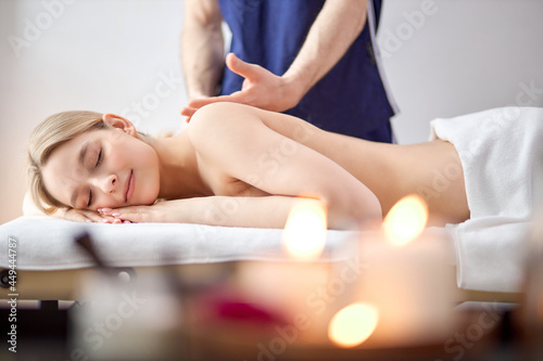 Male hands of massage therapist makes light massage to pretty female in cosmetology room, relaxed shirtless blonde caucasian woman lying on belly getting pleasure, in room with candles