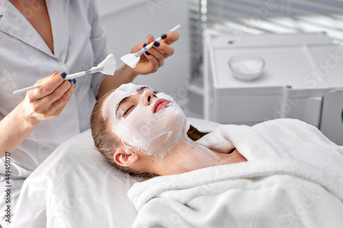 Cropped confident doctor cosmetologist or dermatologist making face mask in cosmetology office. Professional Beautician applying face mask on caucasian woman face lying on bed in bathrobe. copy space