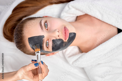 Woman relaxing with charcoal facial mask, cropped professional beautician applying on face of female client, using brush. Top view on female looking at camera, lying on bed in white bathrobe. beauty