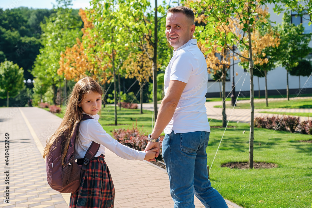 Fototapeta premium Daughter in school uniform and father walking together to school down the street, holding hands, back view