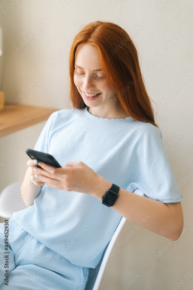 Vertical portrait of laughing young woman wearing casual clothing using mobile phone sitting at desk by window in cozy light cafe. Pretty redhead Caucasian lady having leisure activity in coffee shop. Vertical portrait of laughing young woman wearing casual clothing using mobile phone sitting at desk by window in cozy light cafe. Pretty redhead Caucasian lady having leisure activity in coffee shop.