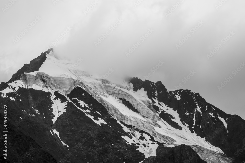 Monochrome atmospheric mountains landscape with big snowy mountain top in low clouds. Awesome minimal scenery with white glacier on black rocks. High mountain pinnacle with snow in clouds in grayscale