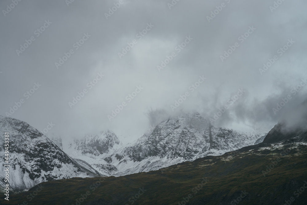 Minimalist gloomy highlands landscape with snow-capped high mountains in overcast weather. Snowbound mountain range in low clouds. Atmospheric minimalism with green mountains, white snow and gray sky.