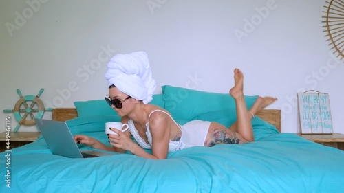 Young beautiful woman with tattoo in white body suit and sunglasses typing on laptop after shower in morning with towel on the head in bed with a cup of coffee. High quality 4k footage