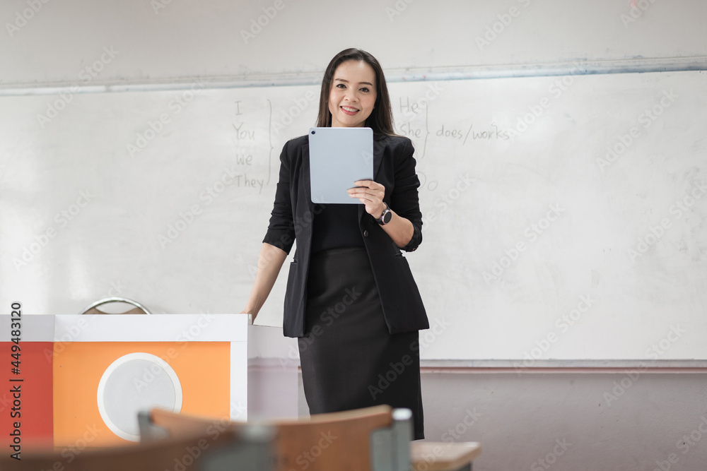 Stock photo portrait of a confident cheerful Asian woman teacher in a black business suit uniform with a digital tablet and laptop to teach modern language in the classroom Stock photo portrait of a confident cheerful Asian woman teacher in a black business suit uniform with a digital tablet and laptop to teach modern language in the classroom