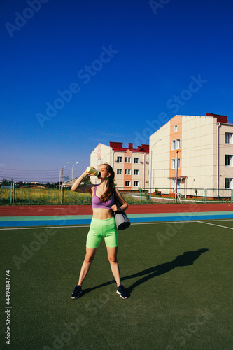 A young woman is standing on the sports field. The girl is holding a rolled-up rug and a bottle of water.