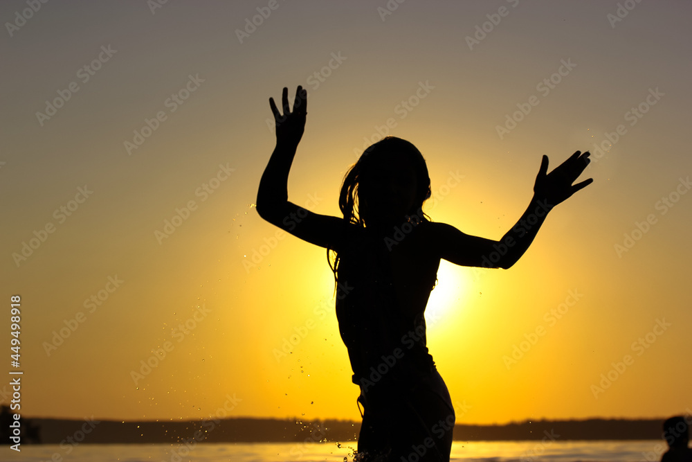 Contrast silhouette of a child teenage girl frolicking in a water body, river, lake, sea, ocean against a yellow sunny sunset sky. Place for text. 
