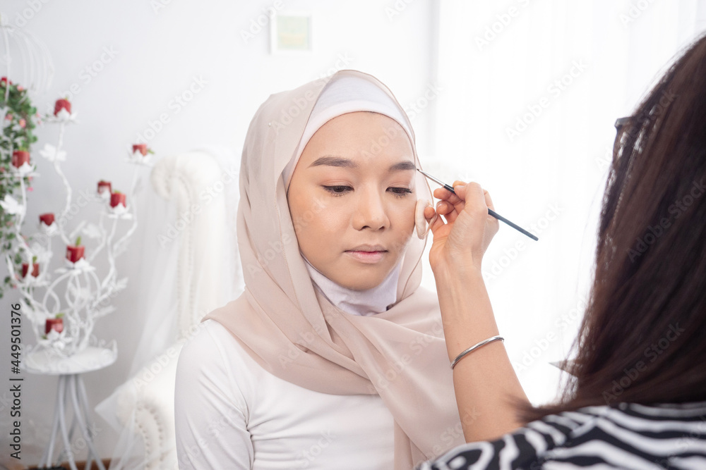 Female makeup artist applying eye brown of young muslim woman in hijab head scarf. Beauty concept.