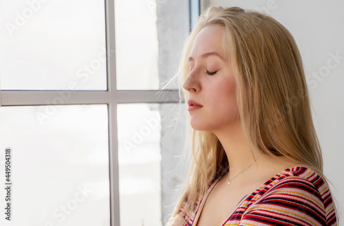 portrait of a young woman with blond hair with closed eyes near the window. Meditating morning process.