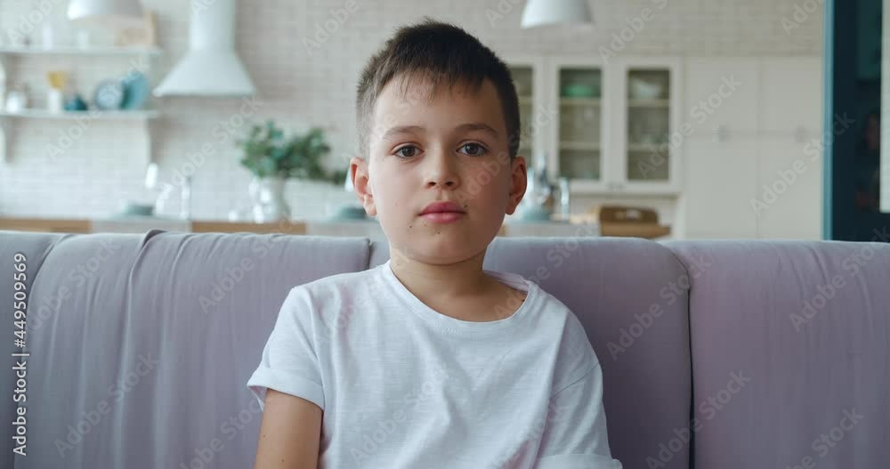 Handsome schoolboy wearing white t-shirt sitting on the sofa in the living room , seriously posing looking at camera.. 4K video footage Handsome schoolboy wearing white t-shirt sitting on the sofa in the living room , seriously posing looking at camera.. 4K video footage