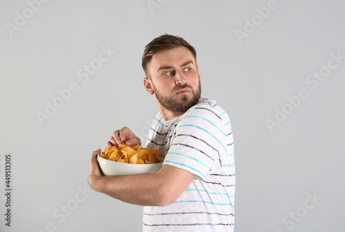 Tableau sur toile Greedy young man hiding bowl with chips on light grey background