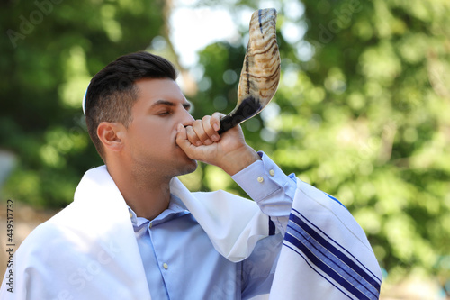 Jewish man in kippah and tallit blowing shofar outdoors. Rosh Hashanah celebration photo