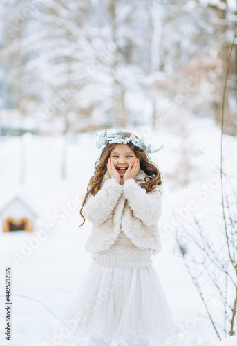 Happy little curly girl in wreath and a white coat playing with snow, in a winter park, standing in the snow, throwing snow. Sunny day. Christmas