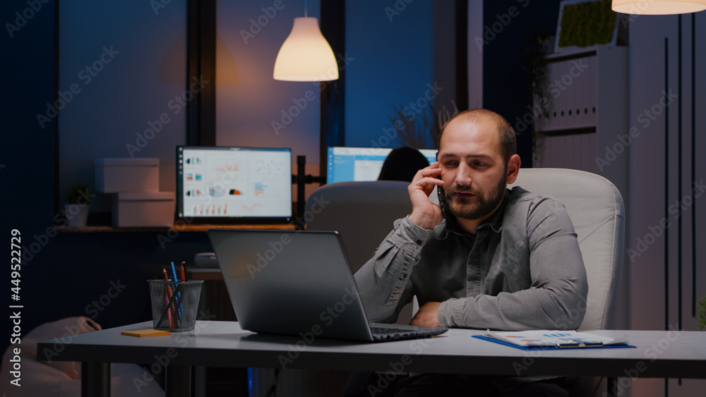 Exhausted businessman sitting at desk table discussing management ideas gesturing during negotiations phone call working late at night in company office. Entrepreneur man planning business interview Exhausted businessman sitting at desk table discussing management ideas gesturing during negotiations phone call working late at night in company office. Entrepreneur man planning business interview