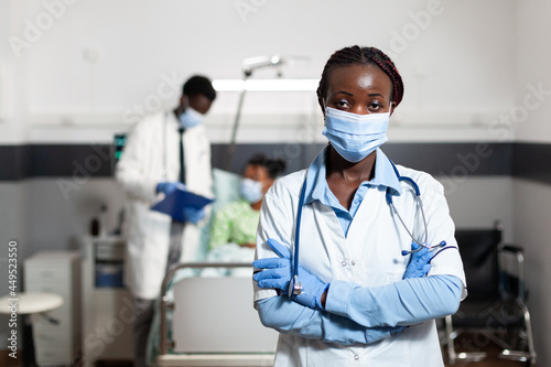 Portrait of african american woman working as doctor in hospital ward clinic. Afro doctor wearing coat  gloves  stethoscope and face mask for protection. Black medic looking at camera