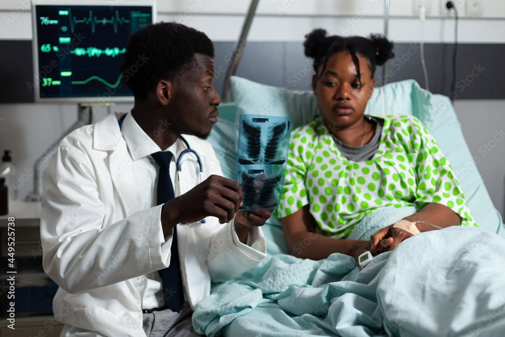 Obraz premium Afro specialist analyzing x ray results with young patient laying in hospital ward bed at clinic. African american doctor showing radiography scan to girl with disease, learning diagnosis
