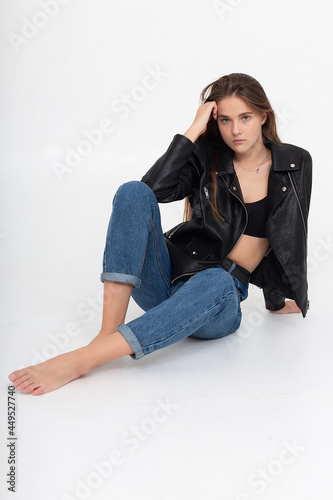 portrait of young caucasian attractive woman with long brown hair in blue jeans and black leather jacket on white background. skinny pretty lady posing at studio with bare feet