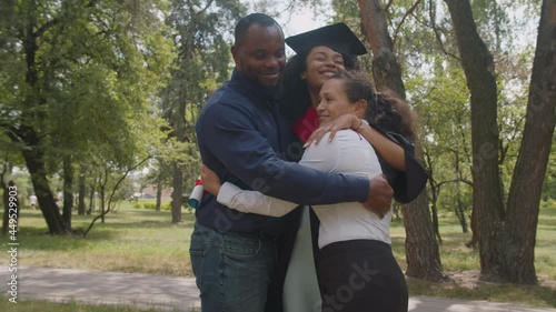 Joyful proud multiracial parents embracing cheerful pretty mixed race female graduate with diploma,dressed in graduation gown and mortarboard, expressing love, care and happiness at graduation day.