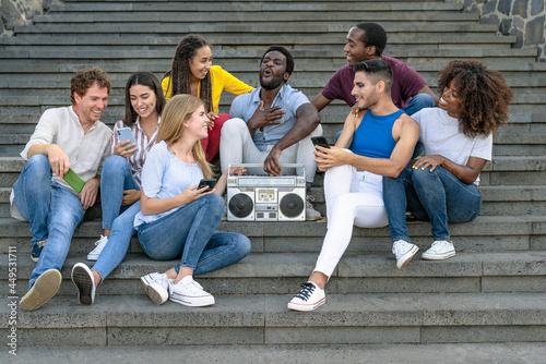 Young multiracial friends having fun listening music with vintage boombox stereo and using mobile smartphone while sitting on urban stairs - Youth millennial lifestyle concept