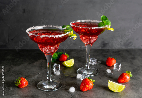 Two glasses of strawberry margarita on the dark table. Lime peel and mint on the glasses. Strawberry berries and lime slices in the background