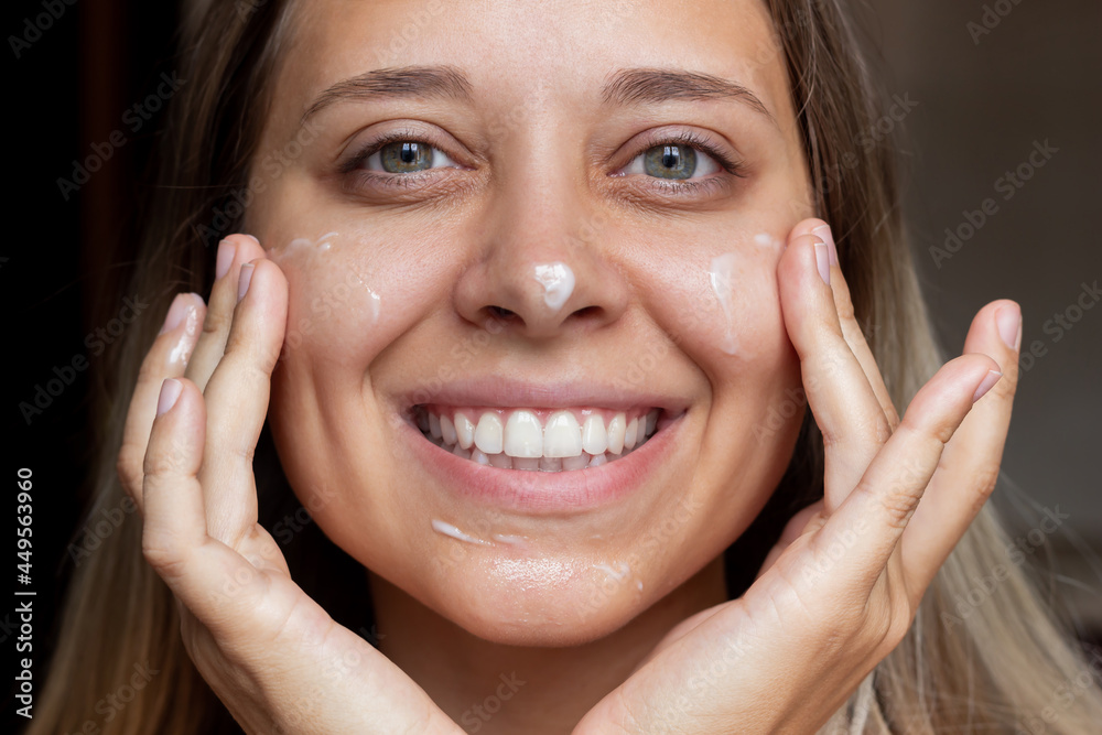 Close-up of a young beautiful smiling caucasian blonde woman applying a moisturizing cream to the skin of the face on a dark background. Skin care, cosmetology Close-up of a young beautiful smiling caucasian blonde woman applying a moisturizing cream to the skin of the face on a dark background. Skin care, cosmetology