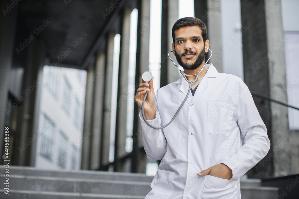 Obraz premium Waist up portrait of smiling pleasant male Indian doctor or medical student in white coat, standing outside hospital or university, demonstrating his stethoscope to camera. Panorama, copy space