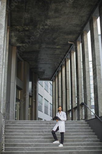 Full length of young handsome skilled male Arab heathcare worker, making notes on folder clipboard, standing on stairs of modern building. Professional Indian doctor filling in medical record outdoors © sofiko14