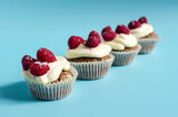 Several cupcakes with white cream and raspberries lined up on a blue background sharpness in the foreground. Sweet homemade cupcakes on a colored background close-up.