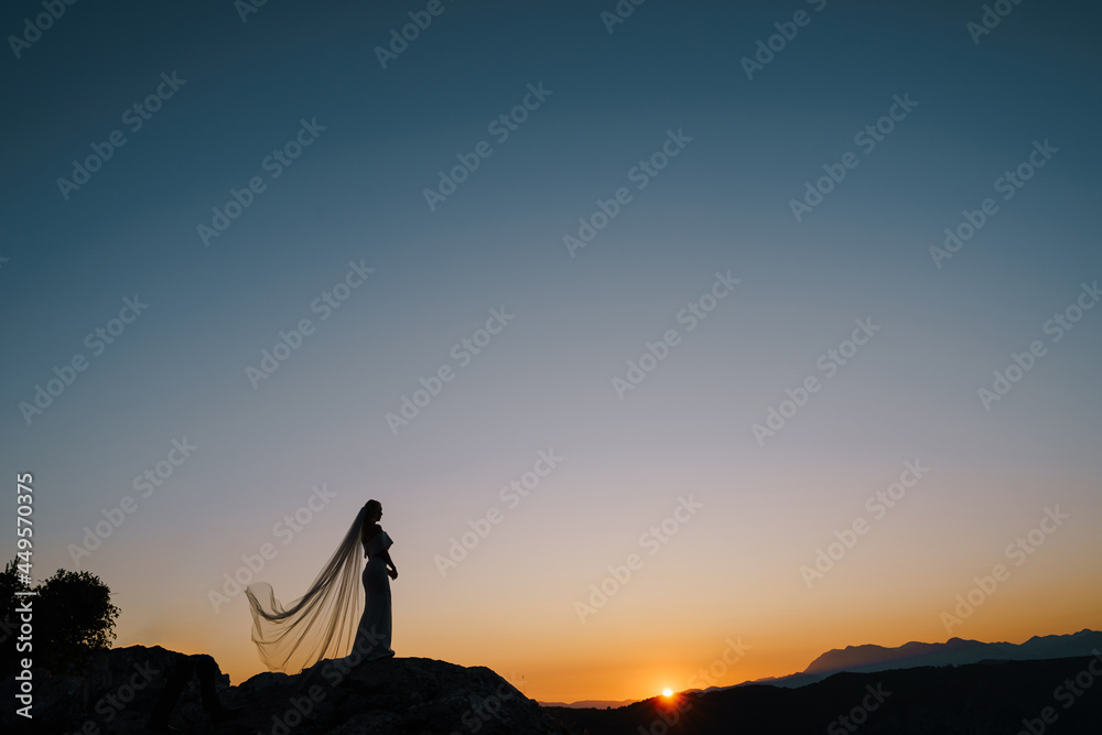 Silhouettes of a bride in a fluttering veil standing on a mountain and looking at the sunset